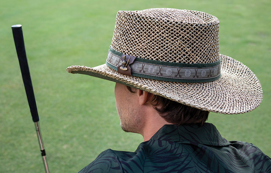 Man wearing a straw hat with golf pins dotted along the green brim standing on a golf course