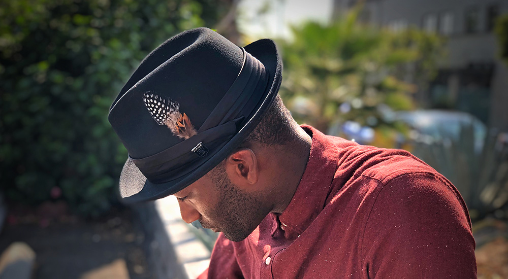 Man wearing a black Stacy Adams hat with orange and black feather in cap and red shirt leaning forward in a backyard of a house