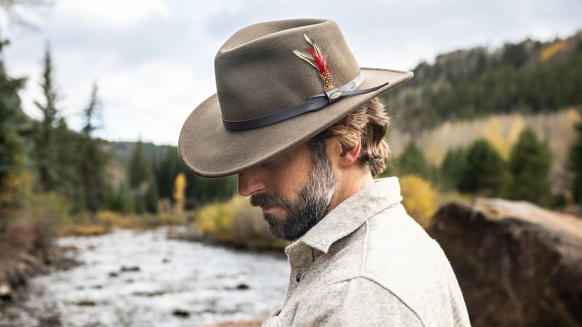 Man wearing a tan hat with red feather and white collared shirt standing in front of a river and forest